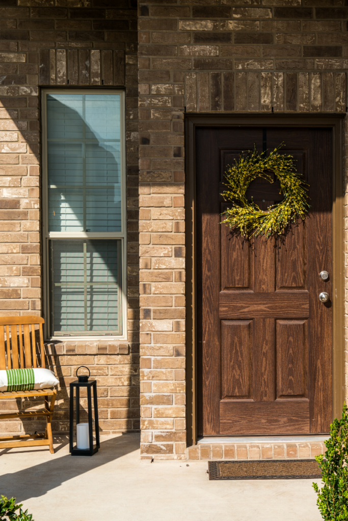 Picture of a home staging front door with wooden door, green wreath, a wooden rocking chair with a green pillow on it with a tall lamp